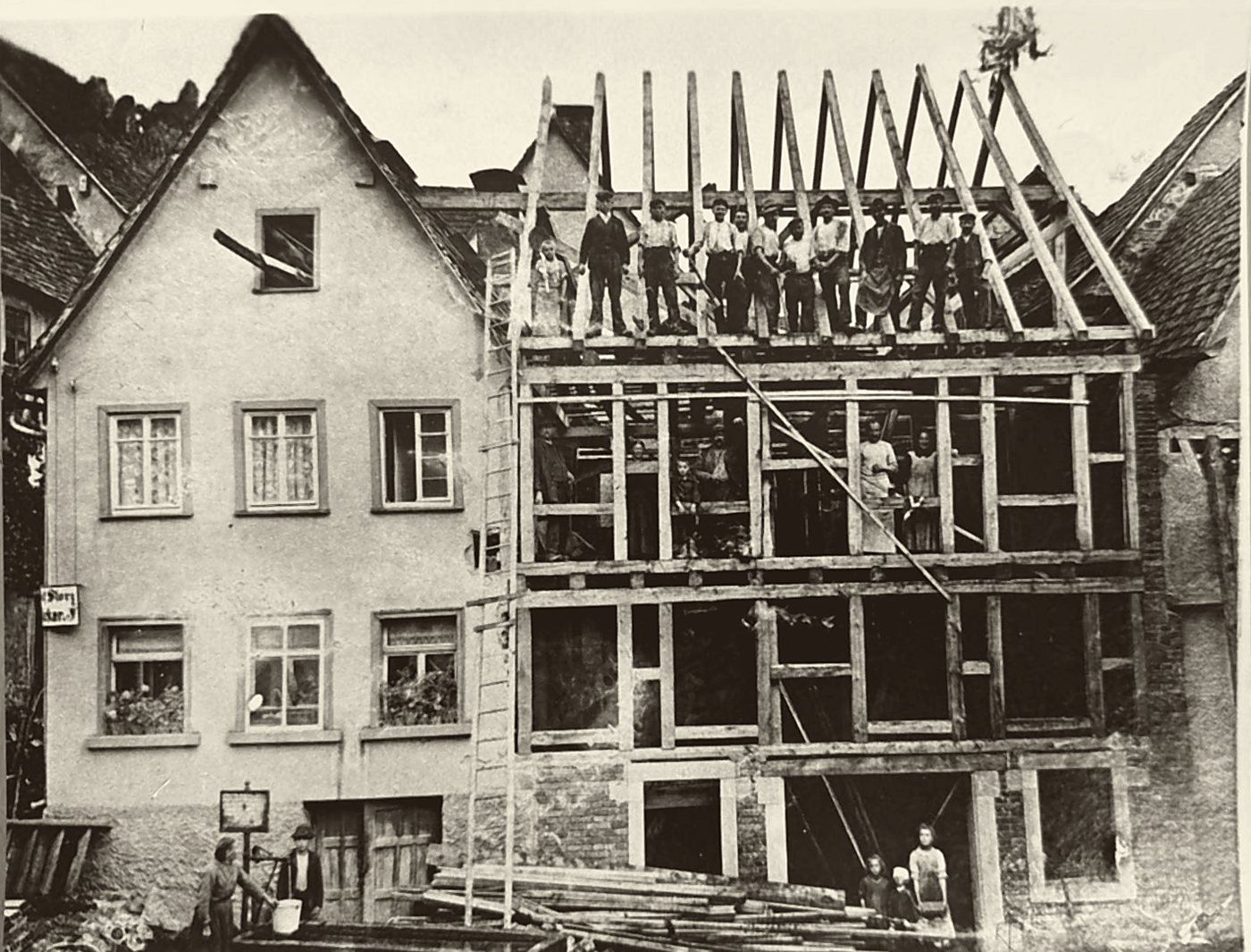 Historische Aufnahme eines Richtfests: Zimmerleute stehen auf den Balken eines neu aufgerichteten Fachwerkbaus, oben hängt der Richtkranz.