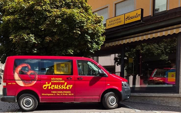 Der Laden von der Straße aus: ockerfarbenes Haus mit gestreifter Markise und dem Leuchtschild Bäckerei Konditorei Heussler, davor der rote Lieferwagen.